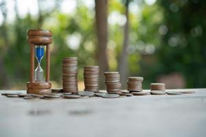 reloj y un montón de monedas de oro sobre la mesa, concepto de tiempo de costo de oportunidad. organice las monedas en montones con las manos, contenido sobre el dinero. la fecha límite y el tiempo es dinero concepto con reloj de arena. foto