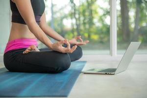 mujer deportiva practicando yoga, haciendo ejercicio ardha padmasana, meditando en posición de loto, haciendo ejercicio, usando ropa deportiva, viendo videos tutoriales de fitness en línea en una laptop, haciendo ejercicio en casa sentada foto
