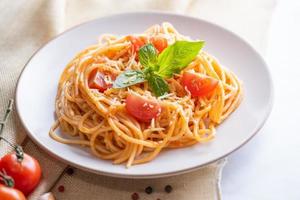 Tasty appetizing classic italian spaghetti pasta with tomato sauce, cheese parmesan and basil on plate and ingredients for cooking pasta on white marble table. photo