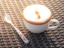 café caliente, capuchino con espuma de leche blanca en una taza de café de cerámica blanca sobre una mesa tejida con bambú hay una pila de café con diseños florales. foto