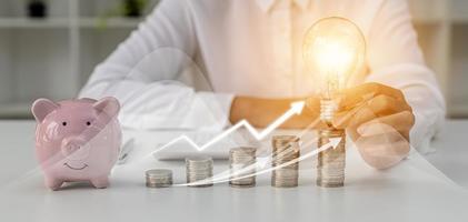 A person holds a light bulb on top of a pile of coins, a large number of coins are placed on the table from low to high, financial chart graphics and growth arrows. personal finance concept. photo