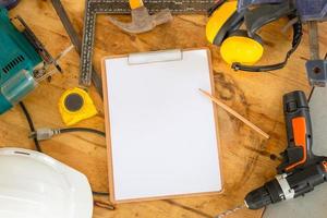 Mockup blank clipboard with white sheet and pencil on carpenter table, White safety helmets and worktools on workbench in woodworking shop photo