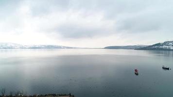 vue sur le lac et les montagnes enneigées en hiver. vue sur le lac en hiver, montagnes enneigées visibles au loin. il y a des bateaux de pêche sur le lac. video