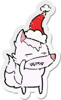 distressed sticker cartoon of a wolf showing teeth wearing santa hat vector