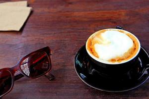 Black cup of hot cappuccino with brown sunglasses and paper towel on the dark brown wooden table. Relaxing time with coffee at cafe shop in the morning photo