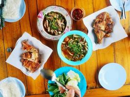 Top view or flat lay of local Thai food with hand and many plates on wood table. Grilled chicken, Sticky rice, Salad, Spicy soup, pork loin and Vegetables. photo