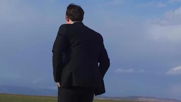 Businessman looking at horizon in field, thoughtful relaxation. Businessman watching the horizon in the field takes his hands out of his pockets and relaxes by lifting them up. video