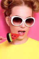 Portrait of stylish cute young blonde woman in sunglasses with colorful watermelon lollipop on pink background photo