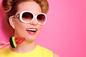 Portrait of stylish cute young blonde woman in sunglasses with colorful watermelon lollipop on pink background photo