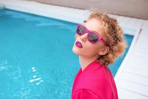 portrait of marvelous stylish redhead curly young woman sitting in fuchsia jacket, stylish sunglasses and black underpants by the swimming pool. photo