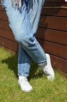 Women's legs in blue jeans and white sneakers on the green grass against the wall. photo