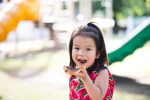 retrato asiático lindo niño comiendo pan con chocolate en el patio de recreo al aire libre. refrigerios para niños cuando juegan y hacen ejercicio hasta que tengan hambre. comida rica. niña de 3 años con vestido rojo. foto