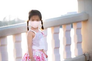 Portrait adorable kid girl wearing cloth face mask travel public park in evening. Summer or spring time. Child stand and watch nature at foot of bridge across pond. Beautiful children aged 5 years old photo