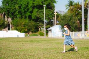 Portrait cute kid girl running on green grass. Child wearing face mask prevent virus and air pollution PM2.5. Children playing sport in summer time. Hot weather. Empty space for enter text. photo