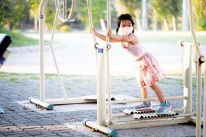 Portrait of adorable kid girl playing treadmill or walking machine is stationary in publics park. Child wearing cloth mask and sneaker.  Exercise and sport time. Summer or spring day. Children 5 years photo