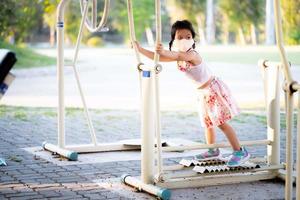 el retrato de una niña adorable jugando a la cinta de correr o a la máquina para caminar está estacionario en el parque público. niño con máscara de tela y zapatillas. tiempo de ejercicio y deporte. día de verano o primavera. niños 5 años foto