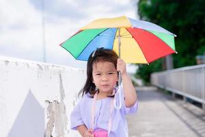 Portrait image of 4-5 years old. Cute Asian girl walks with umbrella in bright sun, to protect her from scorching sunny. In summer time. Kindergarten students are walking home after school. Copy space photo