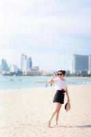 Teenage girl standing on beach looking at beautiful scenery, holding straw hat and sunglasses, smiling brightly. During summer or spring. On comfortable travel day. Empty space. Vertical image. photo