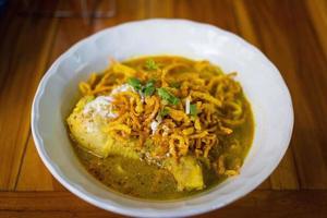 Curried noodle soup with chicken Khao Soi Kai is a Northern Thai food style in white cup on wooden background. photo