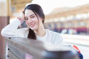 Asian beautiful woman in a long-sleeved white shirt and a hat sits happy smilie in the train station waiting for the train to arrive.