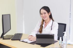 Asian young woman doctor in a white lab coat is sitting on office room at hospital while has a computer, clipbroad and alcohol gel on the deck. photo