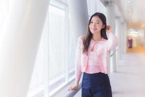 Beautiful teenage Asian girl wears a white with pink shirt and smiles cheerfully while she stands near the glassed window on a nice day. photo