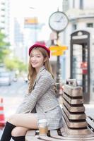 Asian beautiful lady who wears suit and red cap with bronze hair sits on chair in the city outdoors while there was coffee cup on the side on a sunny morning. photo