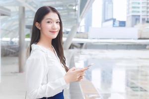 Young Asian business female is going to the office or workplace which she look at camera holds smartphone in her hands in big city with business buildings with the city as a background. photo