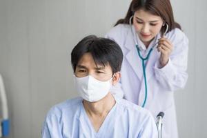 Asian woman doctor use a stethoscope to check the lung rhythm of a male patient who wear face mask while he sit on a wheelchair at hospital in  health care,pollution PM2.5,new normal concept. photo