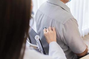 Asian woman doctor wears a surgical mask while she is examining physical of chest by using a stethoscope with a man patient in health care,pollution PM2.5,new normal concept. photo