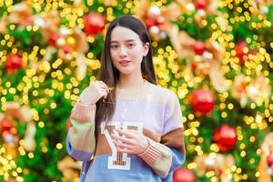 hermosa mujer asiática en un suéter colorido se encuentra feliz. en su mano sostenía una luz frente al árbol de navidad. con bokeh como fondo en el tema de las celebraciones de navidad y año nuevo foto