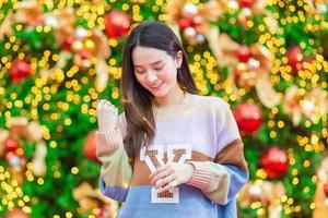 hermosa mujer asiática en un suéter colorido se encuentra feliz. en su mano sostenía una luz frente al árbol de navidad. con bokeh como fondo en el tema de las celebraciones de navidad y año nuevo foto