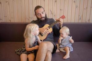 padre barbudo con niños juegan en ukelele interior foto