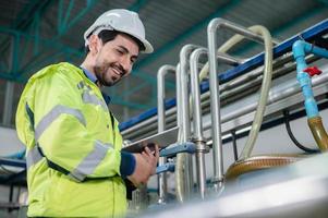 Young male machine inspector wearing vest and hardhat with headphones checking machine and sterilizers in water plant while making notes in digital tablet photo