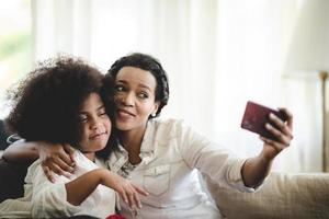 Cheerful young mother taking a selfie using smartphone with sister with curly hair while making funny facial and hand gestures sitting on couch at home photo