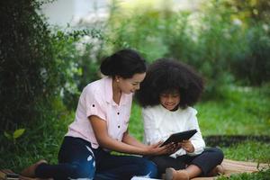 Cheerful young beautiful mother sitting with son with curly hair in park and helping him while using digital tablet while studying photo