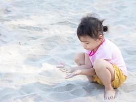 Positive charming 4 years old cute baby Asian girl, little preschooler child playing on sand beach on beautiful sunny day in summer time photo