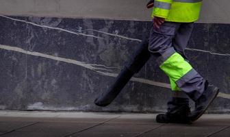 el trabajador de cleseup limpia el suelo de hormigón público con una aspiradora. el trabajador usa uniforme, usa soplador de hojas, limpia la acera en la ciudad. conserje soplando polvo en la acera. ciudad de limpieza para un buen concepto de higiene foto