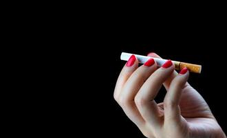 la mano de una mujer sexy con un clavo rojo sostiene un cigarrillo aislado en un fondo negro con espacio para copiar texto. concepto para dejar de fumar. adicto a la nicotina 31 de mayo día mundial sin tabaco. causas del cancer de pulmon foto