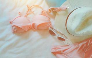 vista superior del traje de baño bikini, gafas de sol y sombrero de paja en la sábana. traje de baño de mujer y accesorios de playa en la cama del resort. Conjunto de bikini rosa para la playa. vacaciones de verano y vibraciones de verano. foto