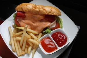 Complete hotdog package. Red sausage, cheese sauce, tomato wedges, lettuce. Plus french fries and a container of red chili and tomato sauce. Served on a white square plate. Close up. top view photo