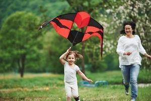 Beautiful background. Positive female child and grandmother running with red and black colored kite in hands outdoors photo