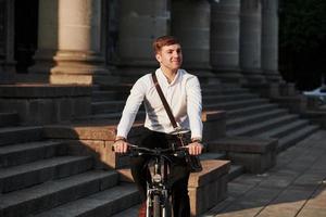 vista frontal. hombre de negocios en ropa formal con bicicleta negra está en la ciudad foto