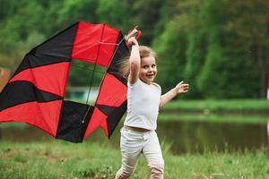 Caucasian ethnicity. Positive female child running with red and black colored kite in hands outdoors photo