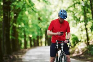 sosteniendo el teléfono inteligente. ciclista en bicicleta está en la carretera asfaltada en el bosque en un día soleado foto