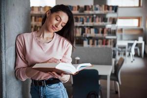 apoyado en la pared. chica morena con ropa informal pasándola bien en la biblioteca llena de libros foto