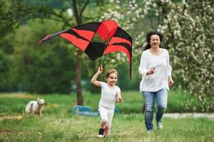 Dog is at background. Positive female child and grandmother running with red and black colored kite in hands outdoors photo