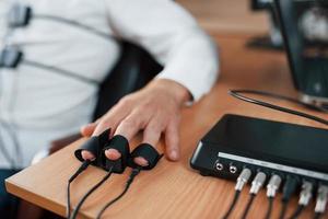 Hand with electrodes on fingers is on the table. Suspicious man passes lie detector in the office. Asking questions. Polygraph test photo