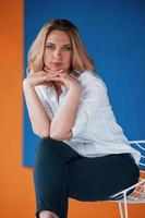 Orange and blue colored wall behind. Businesswoman with curly blonde hair sitting on white chair indoors in room photo