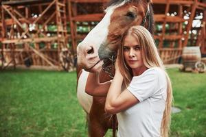 Wooden building behind. Happy woman with her horse on the ranch at daytime photo